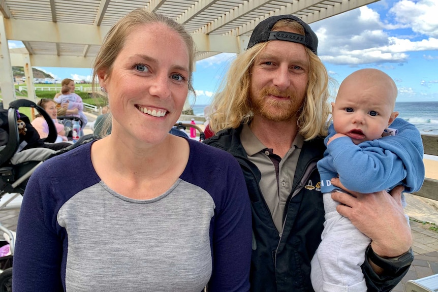 At a beach undercover area, a mother sits next to a father who holds their baby son, with other babies in the background.