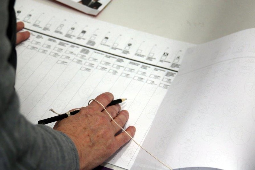 A hand holds a pencil attached to a string at a ballot box. A piece of paper with many boxes, the Senate ballot.