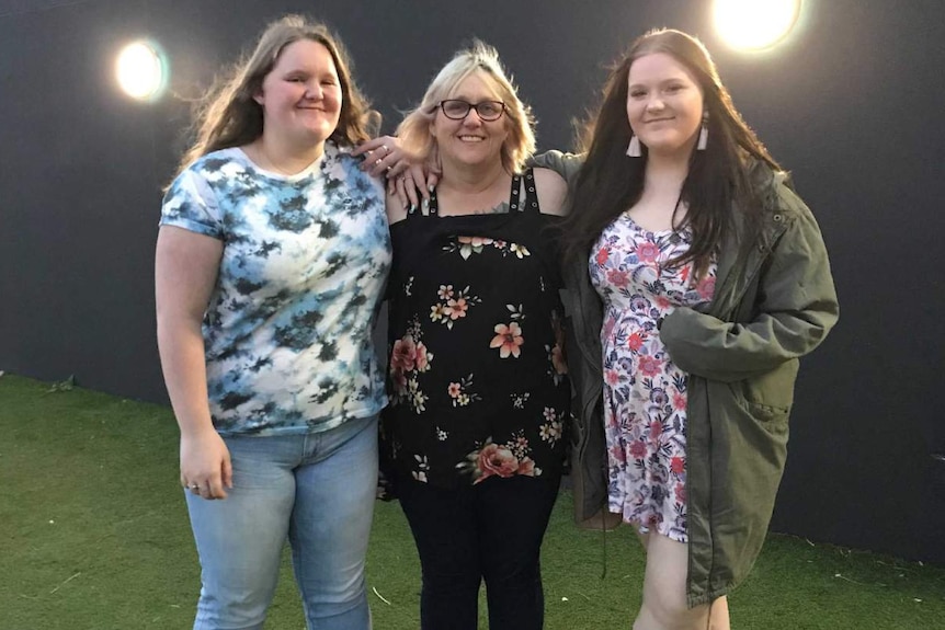 A woman wearing all black and glasses stands with her two daughters on some grass in front of a black building.