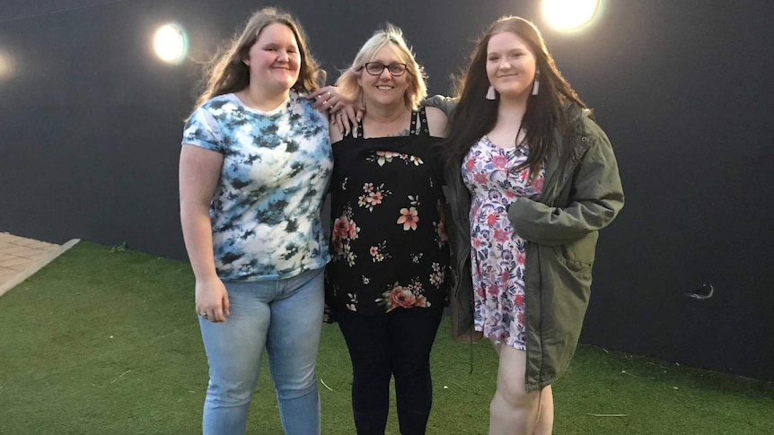 A woman wearing all black and glasses stands with her two daughters on some grass in front of a black building.