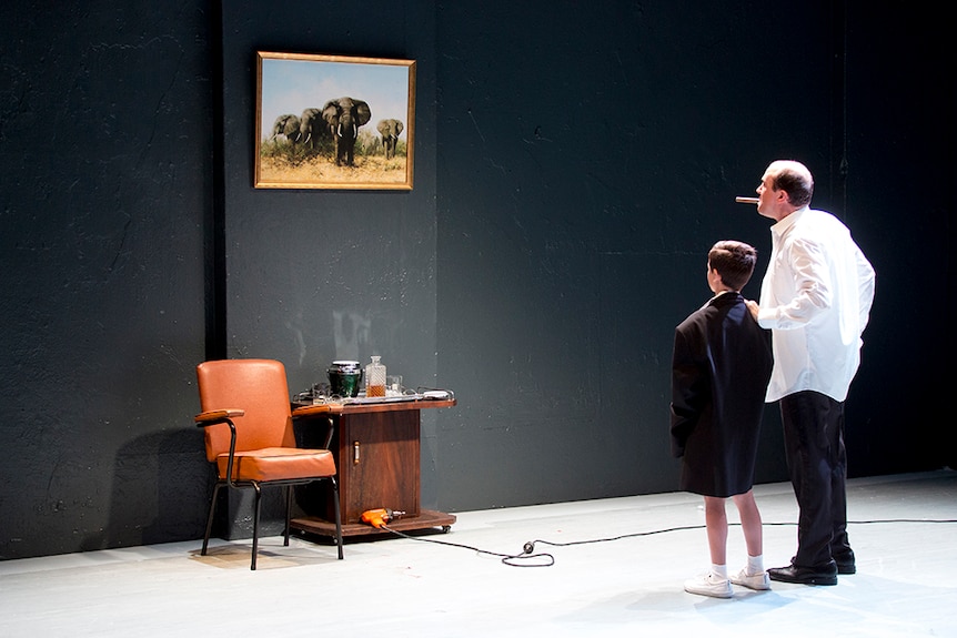 A boy wearing oversized suit jacket stands with an older man cigar, both look at painted elephant artwork on wall on stage.