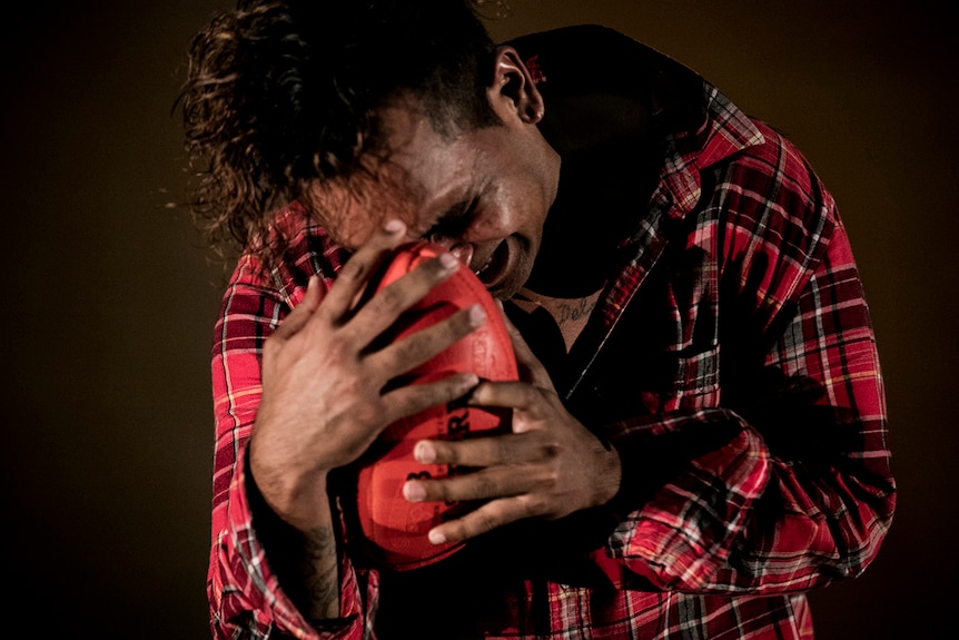 Colour still of Tibian Wyles hunched over crying into a red AFL ball in a production photo for Man with the Iron Neck.
