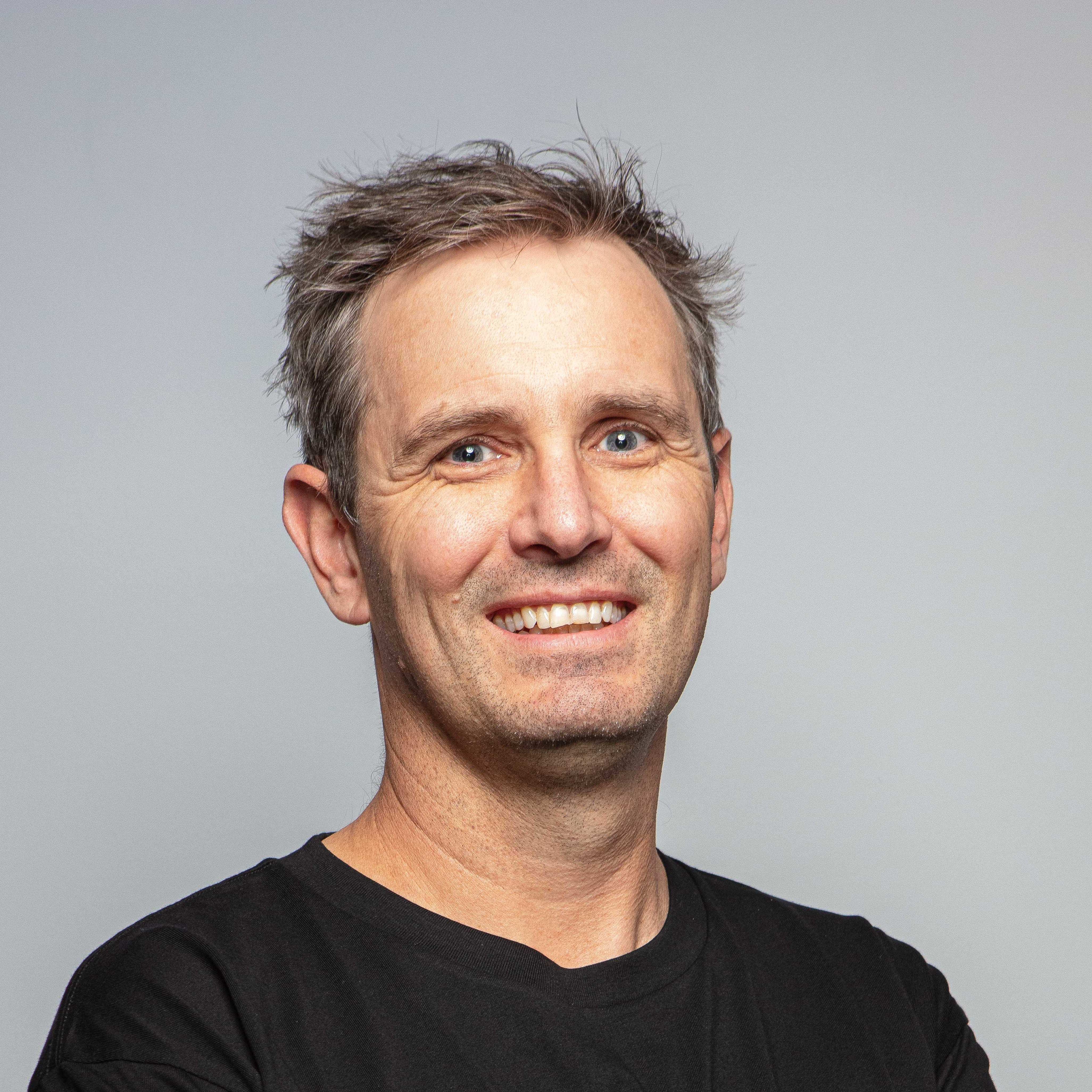A headshot of a white man with browny grey hair, smiling
