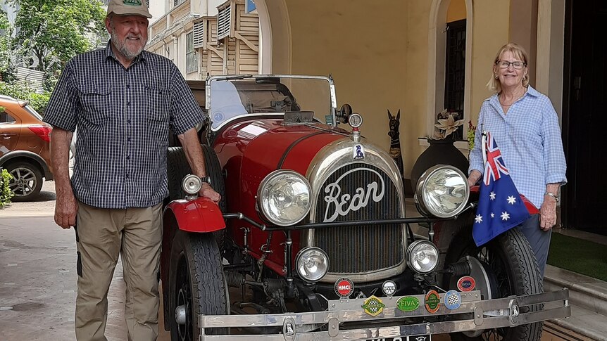 man in cap and grey beard and woman with sandy hair stand either side of red vintage car