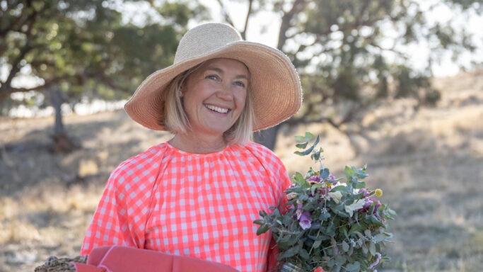 A woman in a bright dress and hat stands holding a picnic blanket and a native bouquet.