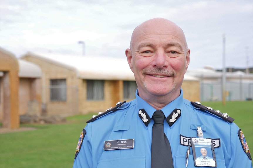 A man in a prison officer uniform stands in front of prison buildings.