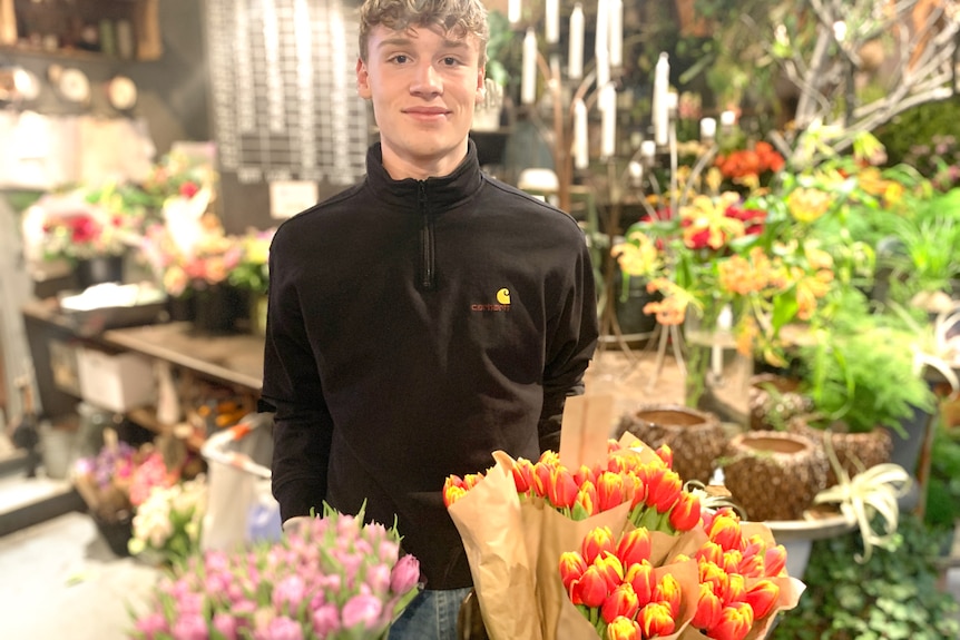 A young man wearing a black shirt stands surrounded by flowers in a shop.