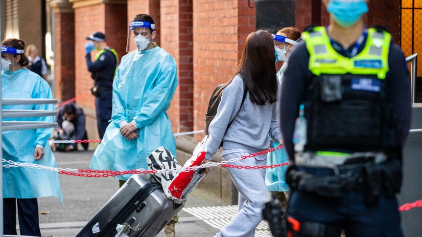 Staff in PPE and police in face masks stand by as travellers tow their luggage past chain barriers at a hotel quarantine site.
