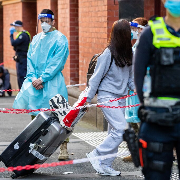 Staff in PPE and police in face masks stand by as travellers tow their luggage past chain barriers at a hotel quarantine site.