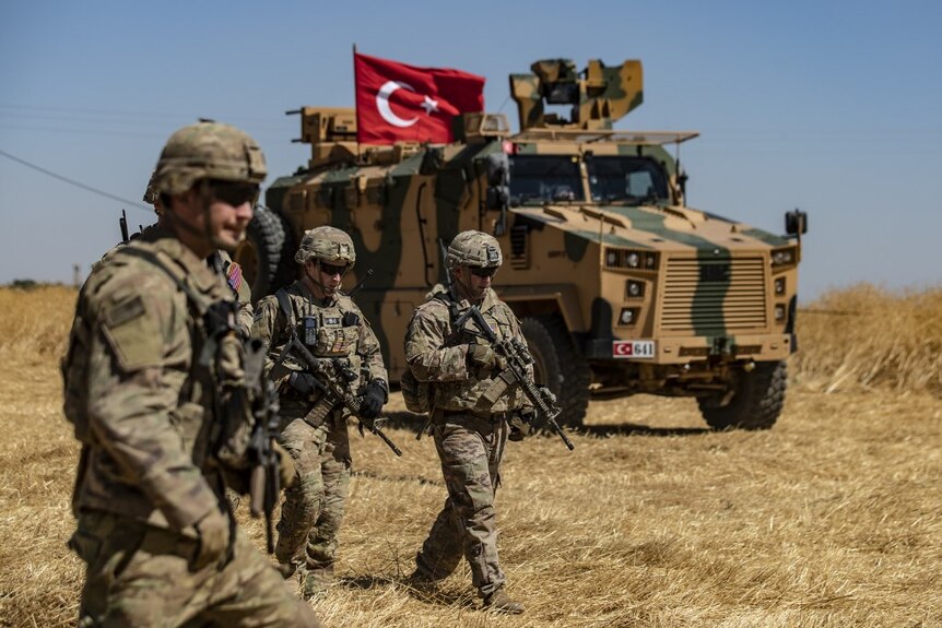 Three armed US troops walk through a field. Behind them is a camouflage Turkish military vehicle which displays a Turkish flag.