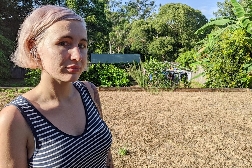 A woman with bobbed hair stands, looking not very impressed, in a yard that has brown grass and no green.