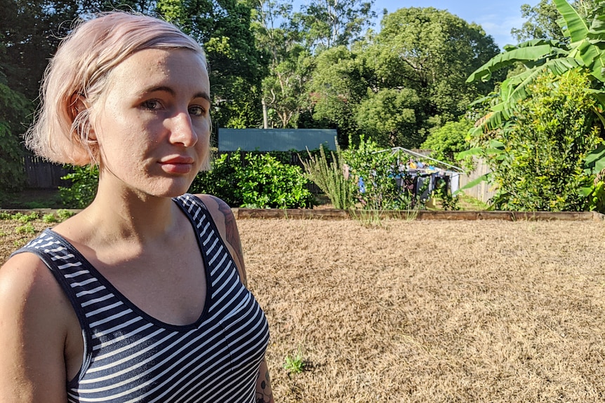 A woman with bobbed hair stands, looking not very impressed, in a yard that has brown grass and no green.