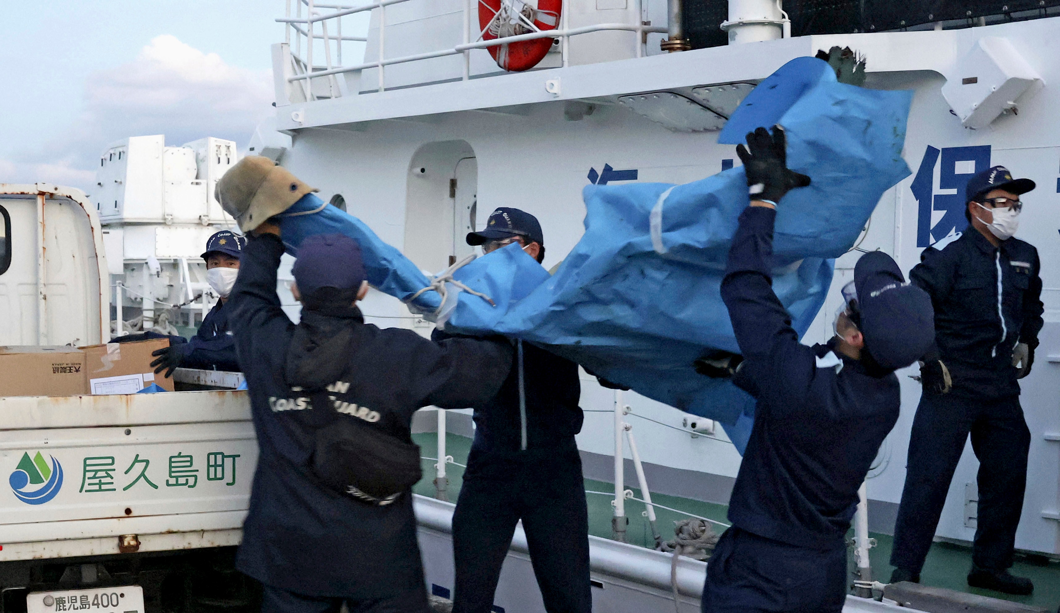 Four men in coast guard uniforms and masks help carry a bag 