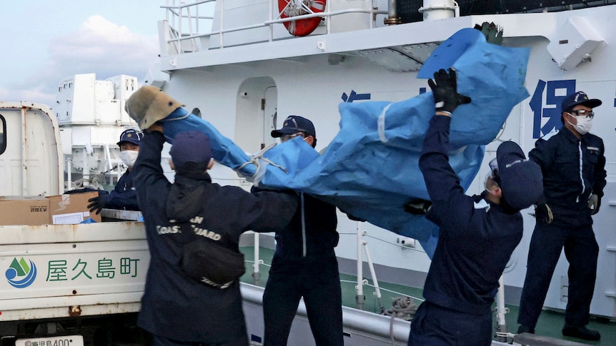 Four men in coast guard uniforms and masks help carry a bag 