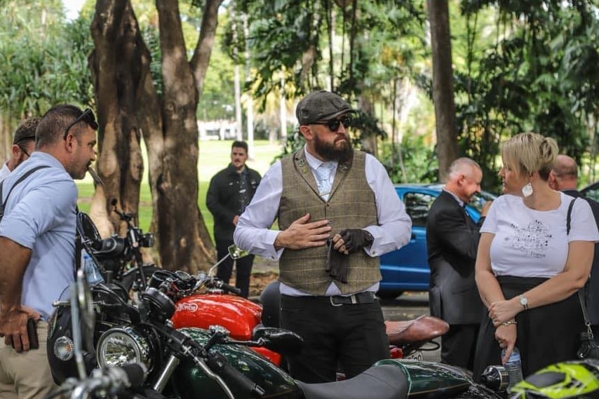 Man wearing a vest and hat standing in front of a classic motorbike talking to people. 