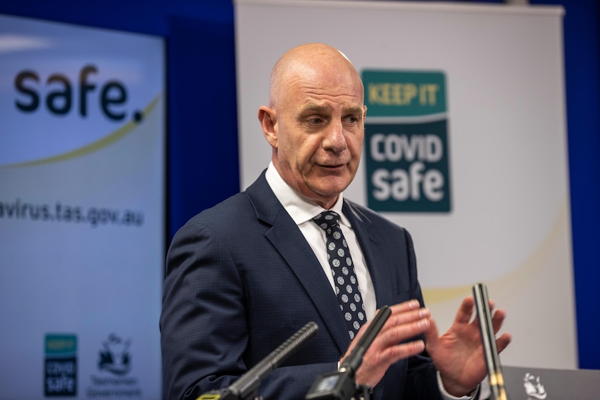 A bald headed man in a suit stands in front of a Covid safe sign