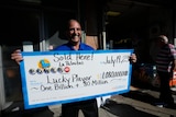 A man with tan skin, a moustache and blue shirt is holding up a large cheque and smiling which says lucky player sold here 