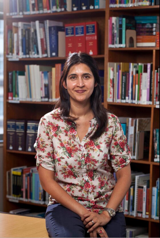 Adila Hassim sits on stool with large bookshelf behind her, with shoulder length brown hair and floral shirt, smiling.