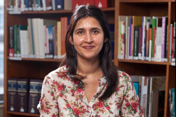 Adila Hassim sits on stool with large bookshelf behind her, with shoulder length brown hair and floral shirt, smiling.
