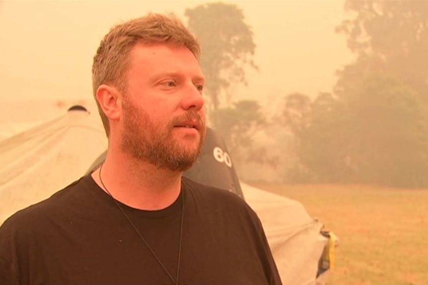 A man with blonde hair stands in a smoke-filled field.