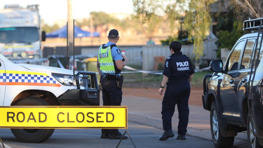 A police officer and a forensic officer stand on a suburban street near two vehicles and a road closed sign, looking away.
