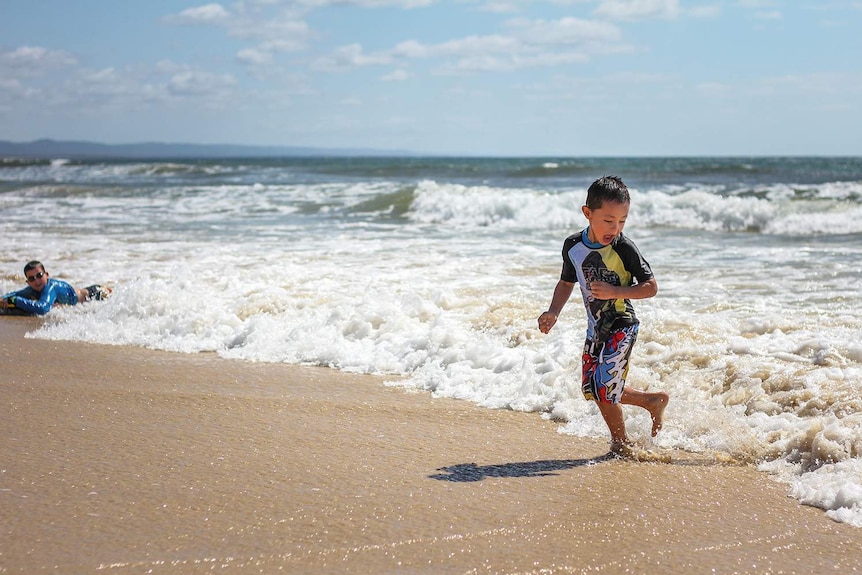 Boy dressed in swimmers runs away from waves while playing at Rainbow Beach on the Fraser Coast.