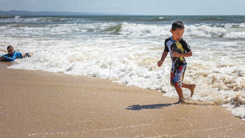 Boy dressed in swimmers runs away from waves while playing at Rainbow Beach on the Fraser Coast.