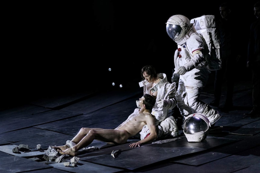 Three figures on black-backed stage, one naked lying in lap of woman, and behind that woman a figure in astronaut suit.