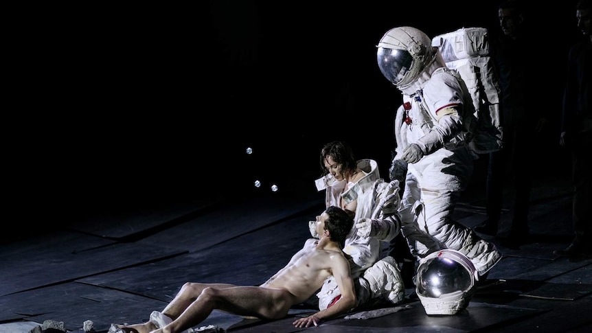 Three figures on black-backed stage, one naked lying in lap of woman, and behind that woman a figure in astronaut suit.