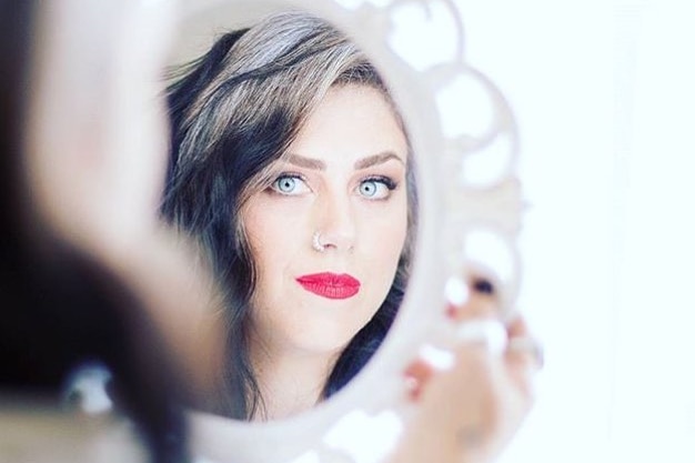 A young woman with grey hair and piercing blue-green eyes looks at her reflection in a handheld mirror