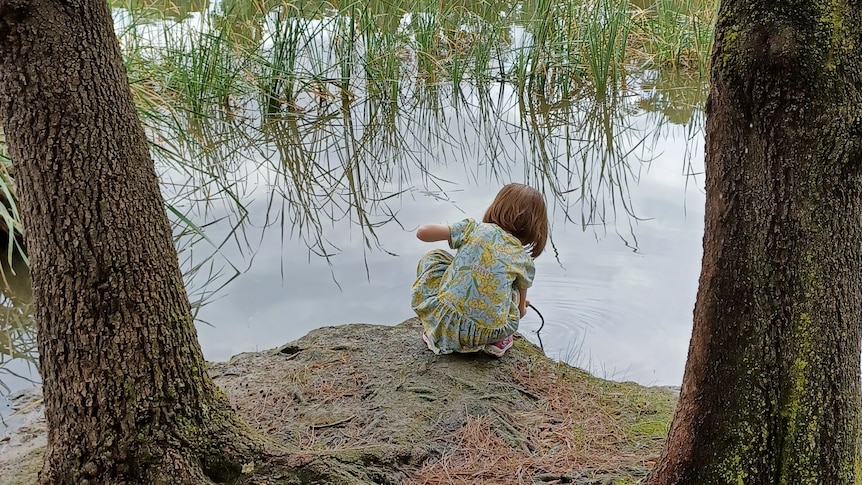 A young child crouches down near the edge of a lake.
