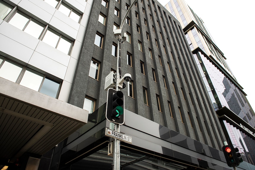 A traffic light with a CCTV camera at its top and city buildings behind it.