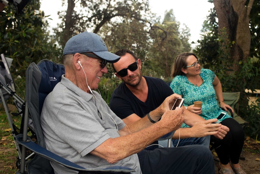 Mike Willesee wearing a cap as he uses headphones to listen to something he's watching on a smartphone in an outdoor setting