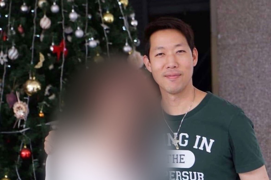 A man with short dark hair stands in front of a Christmas tree with another person whose face is blurred.