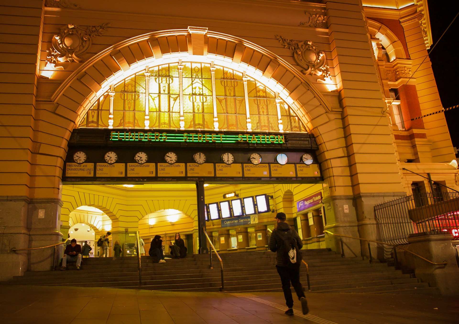 People sit on the steps under the clocks at Flinders Street Station at night.
