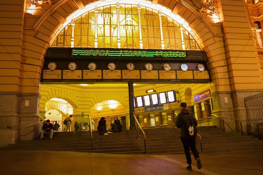 People sit on the steps under the clocks at Flinders Street Station at night.
