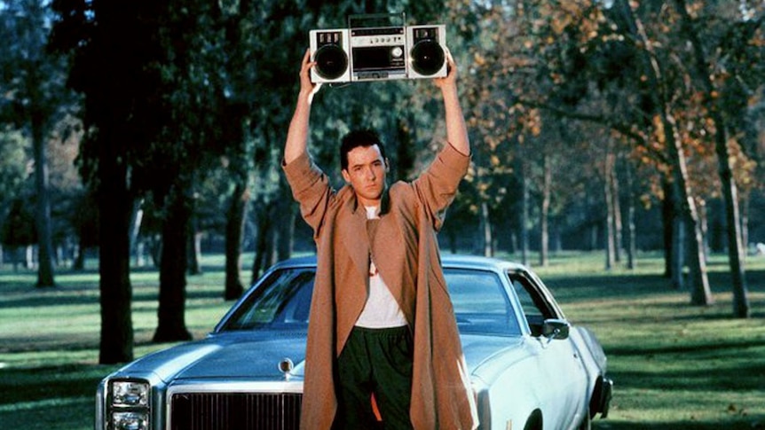 Male in brown trenchcoat holding a boombox above his head, stood in front of blue car
