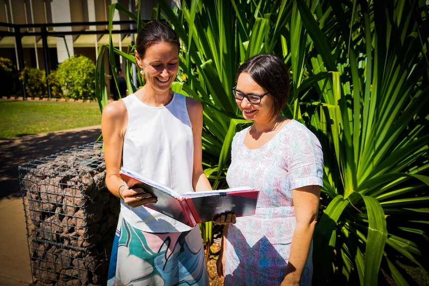 Dr Catherine Marshall and Dr Jane Davies are outside in the green garden. They are looking at an open book and smiling.