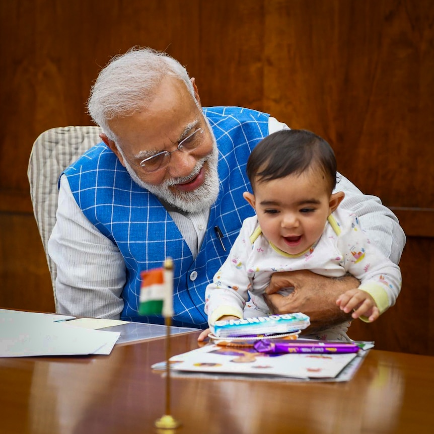 Narendra Modi, wearing a blue checked waistcoat, smiles while holding a baby on his lap at a desk