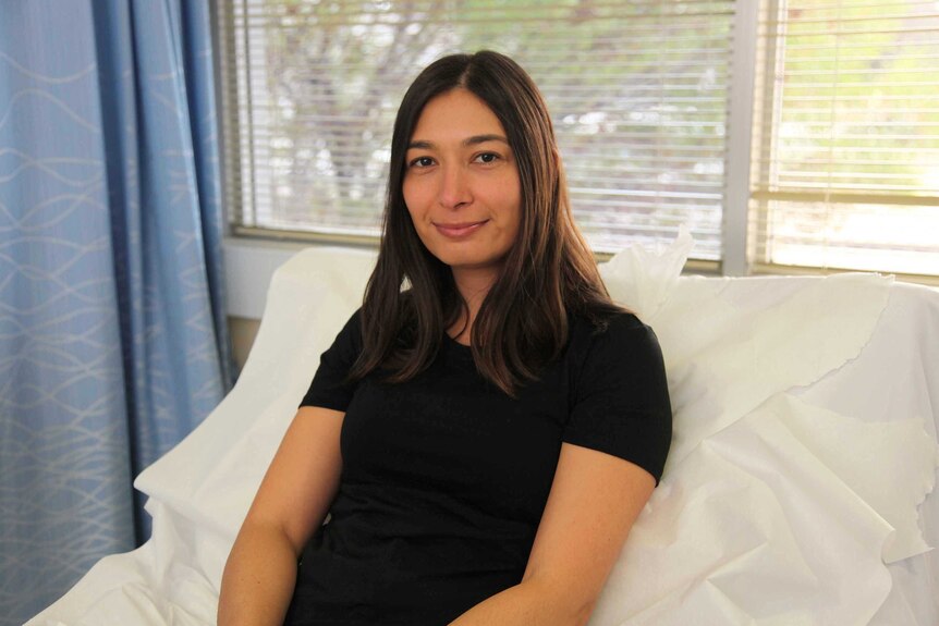 Rachelle Kalic sits on a hospital bed in a black t-shirt waiting to be dosed with a vaccine.