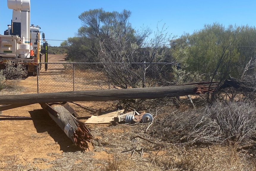 Wooden pole that has been snapped. It is sitting on earth next to bushes with a repair crew and machinery in the backgr