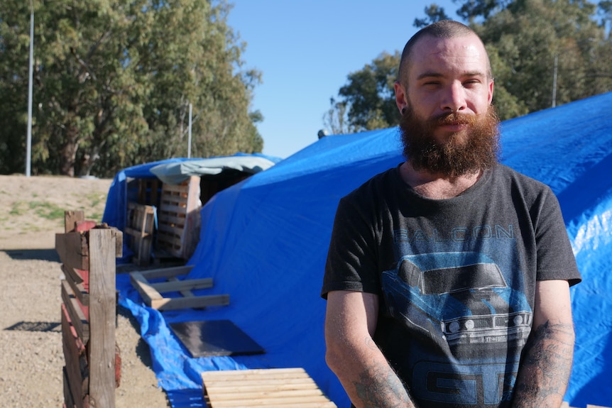 A man with a bushy beard stands in front of pallets propping up tents and a tarp.