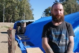 A man with a bushy beard stands in front of pallets propping up tents and a tarp.