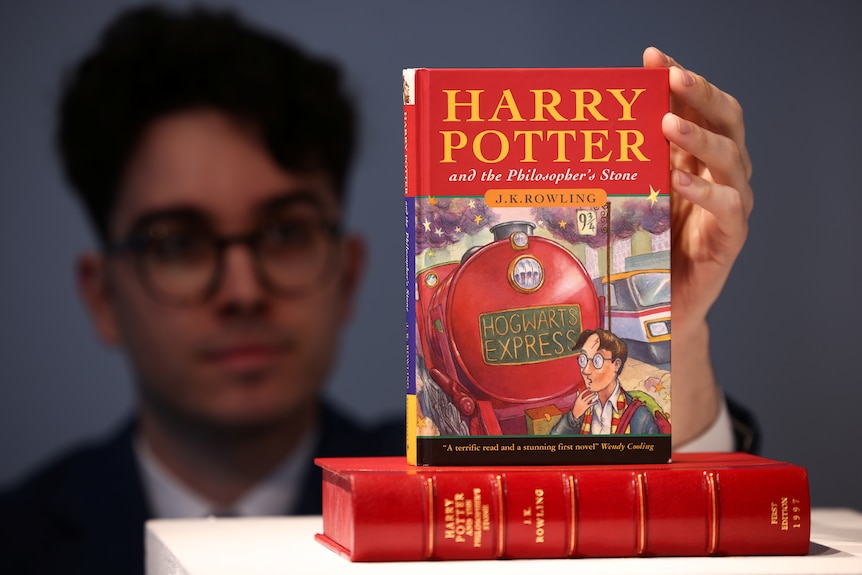 A man with round glasses holds up a rare hardcover edition of Harry Potter and the Philospher's Stone