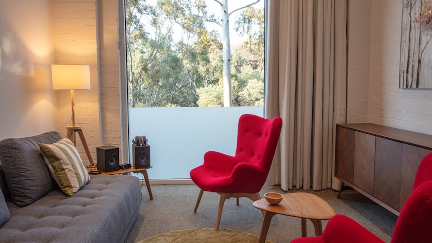 A room with red chairs and a grey sofa, with a large window which looks out onto a canopy of native trees.