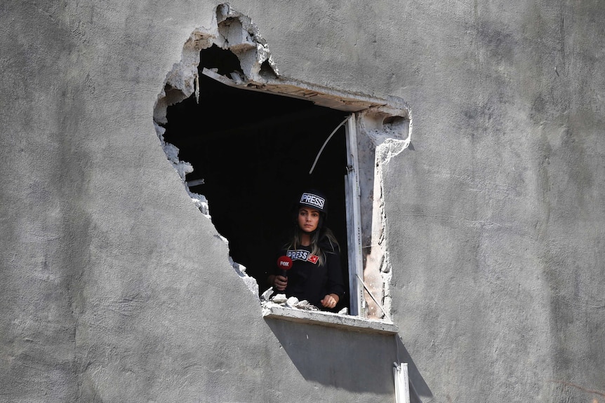 A female journalist looks out of an hole on a house damaged by mortar fire.