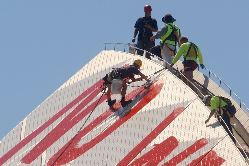 A man with a bucket and hard hat abseils from top of Opera House and paints over red NO WAR letters