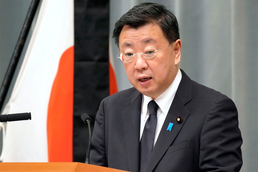 A Japanese MP in a suit stands at a lecturn giving a speech. 