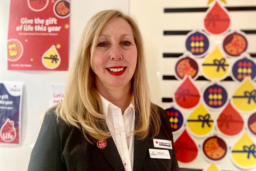 Sally smiles, wearing her Lifeblood badge and standing in front of signs about blood donation.
