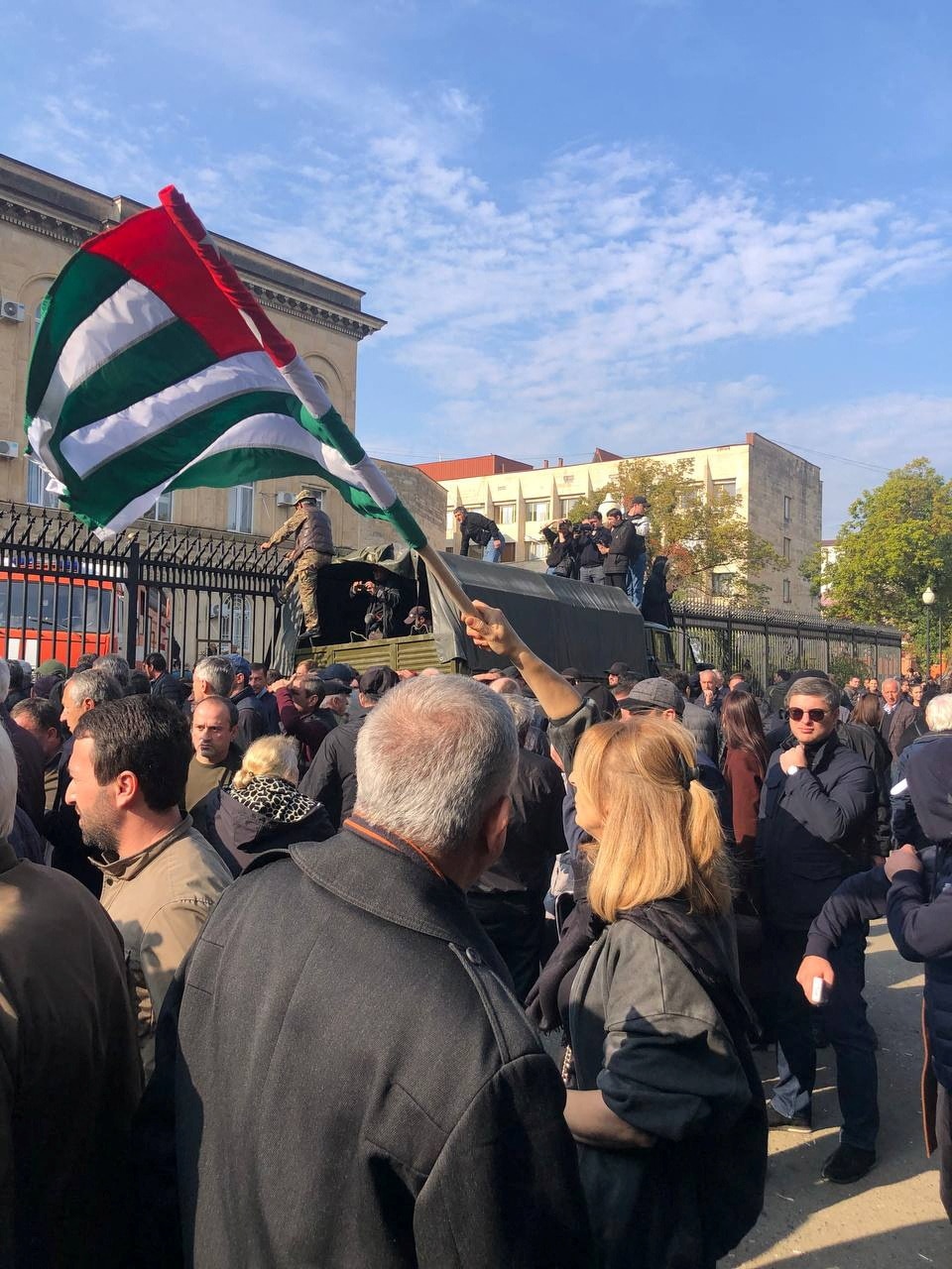 A man waves a flag amid a group of people, back-dropped by a fence and building with guards